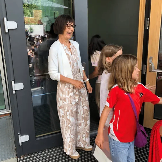 Eine Frau in einem gemusterten Jumpsuit steht in einer Türöffnung mit zwei Mädchen. Das Mädchen rechts trägt ein rotes Shirt mit weißen Akzenten. Bäume und eine Menschenmenge sind durch die Glastür sichtbar.