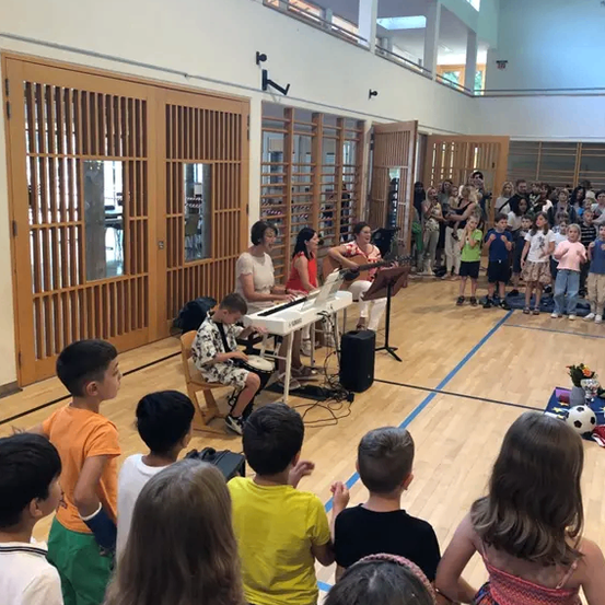 Zwei Frauen spielen Klavier und Gitarre in einer Turnhalle, während eine Gruppe von Kindern aufmerksam zuschaut. Der Raum ist mit Zuschauern gefüllt, und die Umgebung scheint ein Schulereignis zu sein.