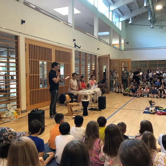 Ein Mann steht vor einem sitzenden Publikum in einem Saal und hält möglicherweise eine Rede. Zwei Frauen sitzen hinter einem Tisch mit Musikinstrumenten. Kinder sitzen auf dem Boden. Es gibt Lautsprecher, einen Fußball und andere Gegenstände auf dem Boden.