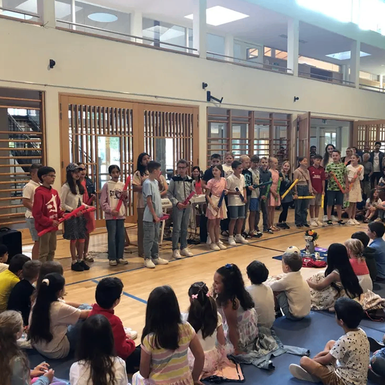 Eine Gruppe von Kindern, einige stehend und einige sitzend, halten Schlagstöcke und scheinen sich in einem Schulflur zu befinden. Sie sind vor einer Lehrerin versammelt.
