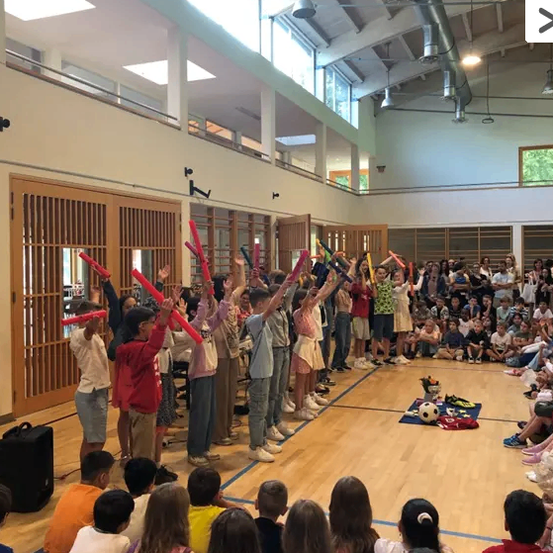 Eine Gruppe von Kindern führt eine synchronisierte Routine mit roten Stöcken in einer Turnhalle durch. Viele Erwachsene schauen zu.