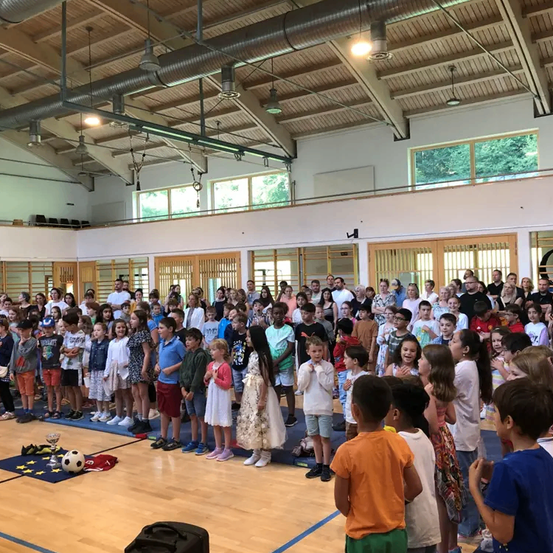 In einer Turnhalle versammeln sich eine große Gruppe von Kindern und Erwachsenen. Sie stehen in Reihen, manche mit gefalteten Händen. Ein Fußball und ein Pokal liegen auf dem Boden. Die Turnhalle hat Holzböden, Glasfenster und hängende Lampen.