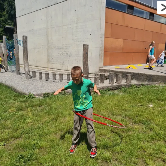 Ein Junge schwingt einen roten Hula-Hoop in einem Grasbereich in der Nähe eines Gebäudes. Hinter ihm gehen zwei Mädchen auf einem Betonweg. Bäume und ein Zaun sind im Hintergrund.