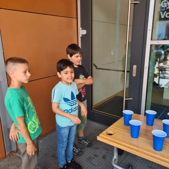 Drei Jungen stehen vor einer Tür mit einem Tisch, auf dem blaue Becher stehen. Der Junge in der Mitte trägt ein hellblaues Adidas-Shirt.