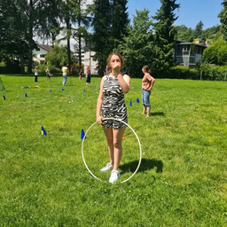 Ein Mädchen steht in einem Park mit einem Hula-Hoop, während andere im Hintergrund spielen. Bäume und Gebäude sind in der Ferne sichtbar.