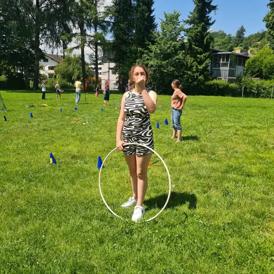 Ein Mädchen steht in einem Park mit einem Hula-Hoop, während andere im Hintergrund spielen. Bäume und Gebäude sind in der Ferne sichtbar.