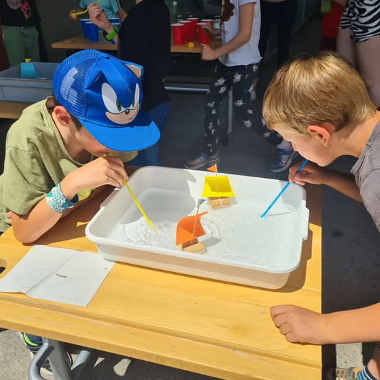 Zwei Jungen, einer mit einer Sonic-Kappe, treten in einem Wassertablett gegen Miniaturboote an. Einer hält einen Strohhalm und pustet in das Wasser.