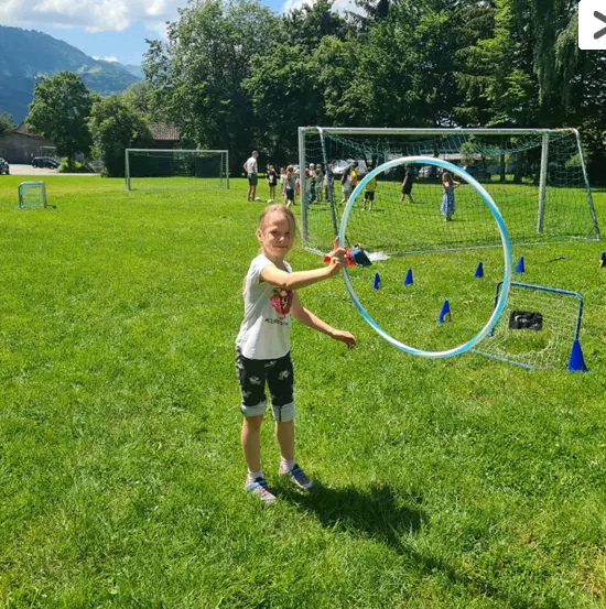 Ein Mädchen wirft einen Wasserballon auf ein Ziel an einem sonnigen Tag auf einem Rasenfeld mit Toren im Hintergrund.