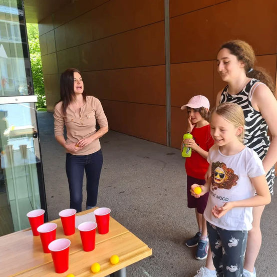 Drei Frauen und zwei Mädchen spielen Bierpong auf einem Holztisch mit roten Bechern und gelben Bällen. Sie stehen draußen in der Nähe einer Glastür.