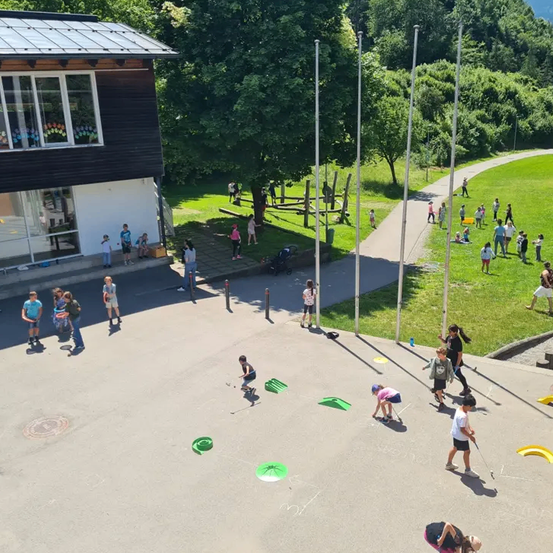 Kinder spielen auf einem Spielplatz mit einem Gebäude im Hintergrund. Einige stehen, andere sind gebeugt, wahrscheinlich spielen sie ein Spiel. Pfosten sind auf dem Boden verstreut.