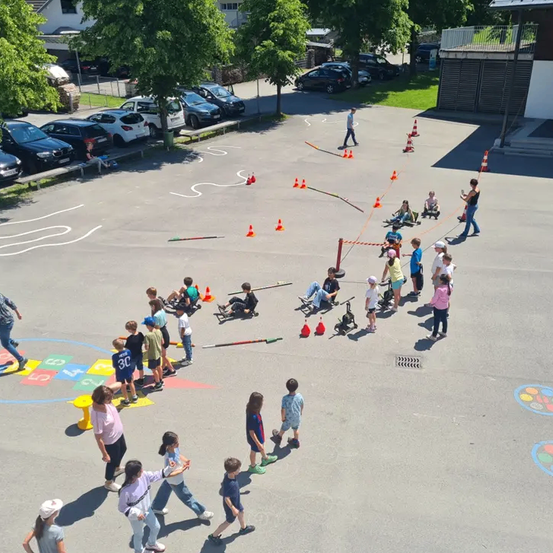 Eine Vogelperspektive eines Spielplatzes mit Kindern, die verschiedene Spiele spielen. Sie laufen, sitzen und stehen auf bemalten Mustern und um orangefarbene Kegel herum. Ein Mann geht an der Seite.