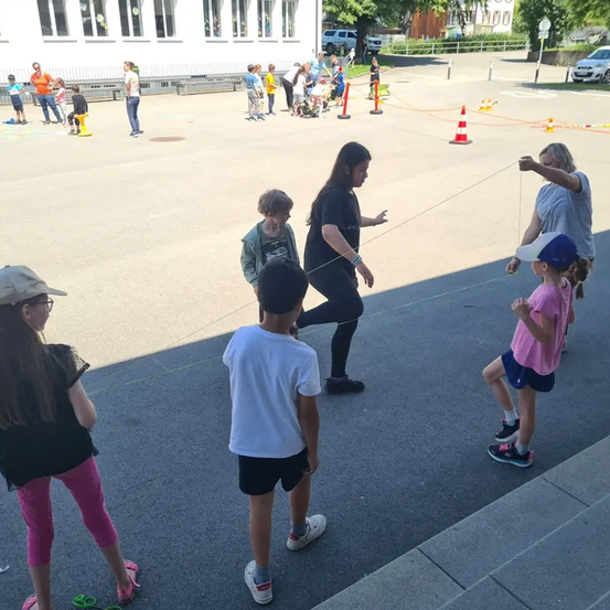 Eine Gruppe von Kindern und Erwachsenen befindet sich auf einem Spielplatz mit Verkehrskegeln und einem Gebäude im Hintergrund. Eine Frau in einem schwarzen Shirt zieht an einer Schnur.