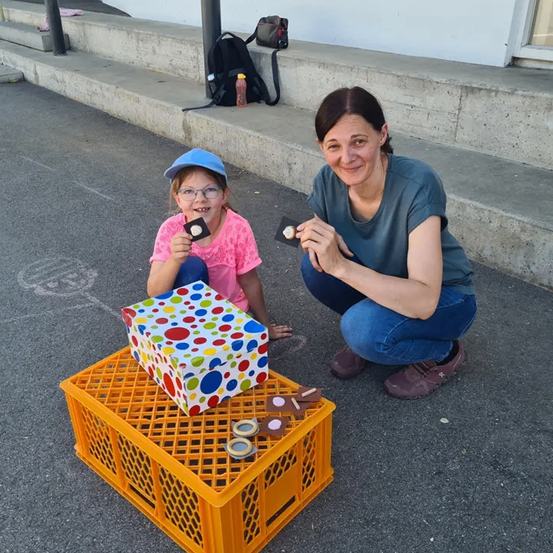 Eine Frau und ein Mädchen sitzen auf der Straße mit einer Schachtel mit Süßigkeiten, beide halten ein Stück. Die Schachtel hat farbige Punkte. Ein Rucksack und eine Flasche sind auf den Stufen.