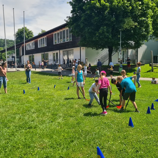 Eine Gruppe von Kindern und Erwachsenen spielt auf einem Rasenfeld mit verschiedenen Spielen und Aktivitäten. Einige benutzen Stöcke und Reifen.