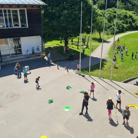 Eine Gruppe von Kindern spielt in einem Außenbereich mit einem Gebäude, einem Weg und einem Rasenfeld. Einige Kinder spielen Golf mit grünen Frisbees auf dem Boden.
