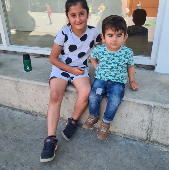 Zwei Kinder sitzen auf einer Betontreppe. Ein Mädchen in gepunkteter Shorts und Turnschuhen, und ein Junge in Jeans und einem grünen Shirt. Ein grüner Thermosbecher steht hinter ihnen. Ein Gebäude mit Glasfenstern und Reflexionen von Menschen draußen.