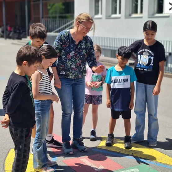 Eine Frau erklärt etwas einer Gruppe von Kindern, die auf einem nummerierten Spielplatz stehen. Sie stehen vor einem Gebäude.