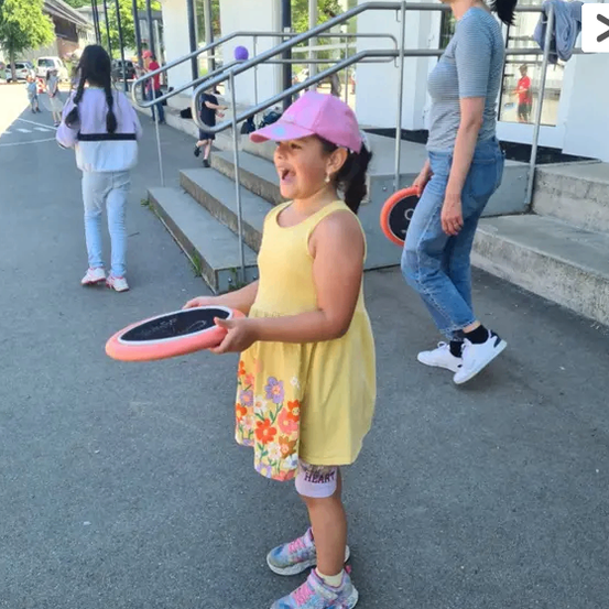 Ein Mädchen in einem gelben Kleid hält einen rosa Frisbee und steht in einem Außenbereich mit einer Frau hinter ihr und einem Gebäude mit Treppen.