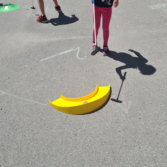 Ein kleines Mädchen mit einem Golfschläger zielt auf ein gelbes Hindernis auf dem Boden. Andere Menschen spielen ebenfalls auf dem Boden.