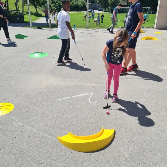 Drei Kinder spielen Minigolf auf einem gepflasterten Außenplatz. Es gibt gelbe und grüne Hindernisse auf dem Boden. Im Hintergrund ist eine Wiese mit Bäumen und Bergen zu sehen.