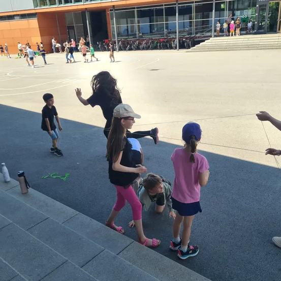 Eine Gruppe von Kindern spielt auf einem Schulhof, wobei einige Erwachsene beaufsichtigen. Ein Kind kriecht auf dem Boden, während ein anderes ein Seil hält.