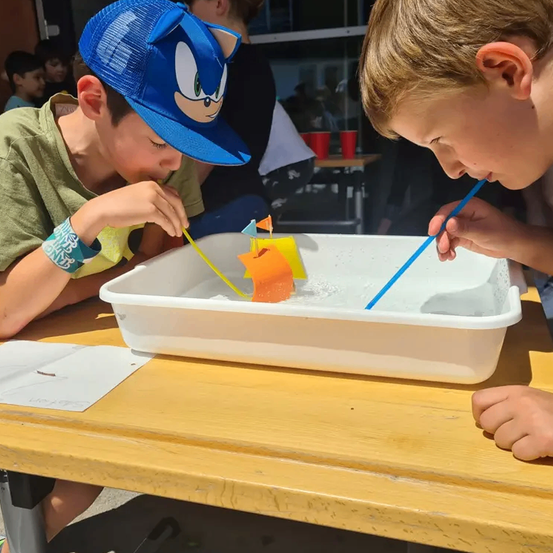 Zwei Jungen sitzen an einem Tisch und blasen in Strohhalme, mit einem kleinen Boot, das in einer Wasserschale vor ihnen schwimmt. Ein Junge trägt eine Sonic the Hedgehog-Kappe.