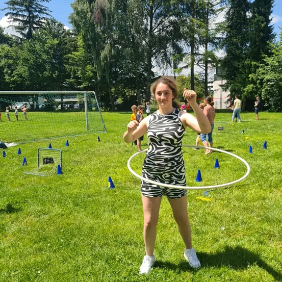 Eine Frau steht auf einem Rasenfeld, hält einen Hula-Hoop und hat ein Fußballtor und blaue Kegel im Hintergrund. Bäume und ein Gebäude sind in der Ferne.