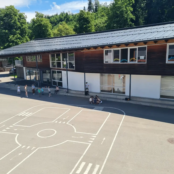 Ein Gebäude mit einem Metalldach steht in einem Park mit Bäumen. Menschen gehen oder sitzen davor. Ein weißes Diagramm ist auf dem Boden gemalt.