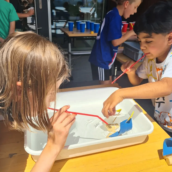 Kinder spielen mit Booten in einer Wasserwanne auf einem Tisch und benutzen rote Strohhalme. Ein Junge und ein Mädchen konzentrieren sich auf ihre Boote. Im Hintergrund stehen andere Kinder um einen Tisch mit Bechern.