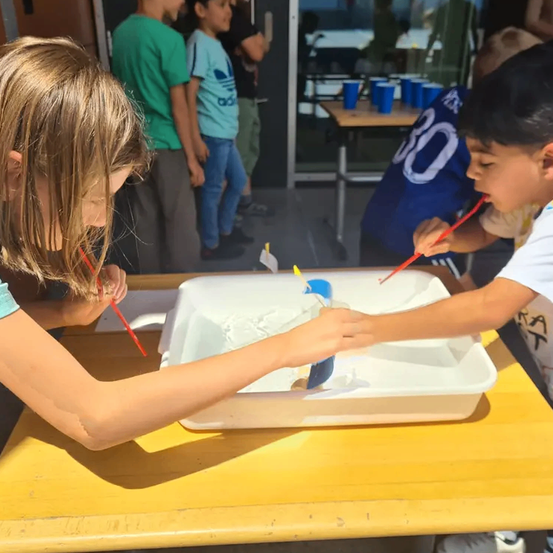 Ein Mädchen und ein Junge sind in eine kreative Aktivität vertieft und mischen und spielen mit weißem Sand unter Verwendung roter Pinseln. Sie sitzen an einem Holztisch, während andere Kinder im Hintergrund zu sehen sind.