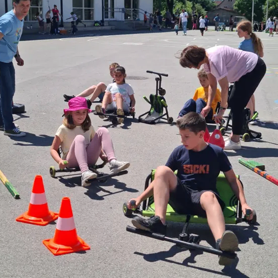 Eine Gruppe von Kindern fährt auf Go-Karts auf einem gepflasterten Außenbereich. Einige Kinder sitzen auf den Go-Karts, während andere stehen. Eine Frau steht in der Nähe der Go-Karts, und Verkehrskegel sind auf dem Boden platziert. Ein Gebäude ist im Hintergrund.