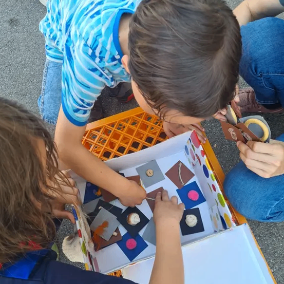 Drei Kinder sitzen auf dem Boden und erkunden eine offene Box mit verschiedenen farbigen Gegenständen darin, möglicherweise in einer Sortieraktivität beschäftigt.