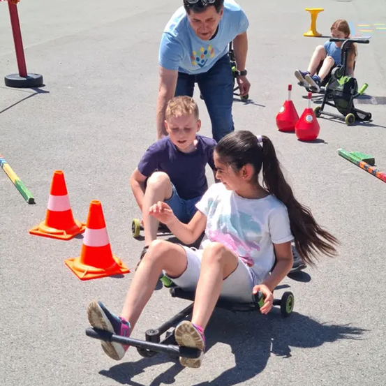 Ein Mann hilft zwei Kindern, die Go-Karts auf einer asphaltierten Straße fahren, umgeben von Verkehrskegeln und Menschen im Hintergrund.