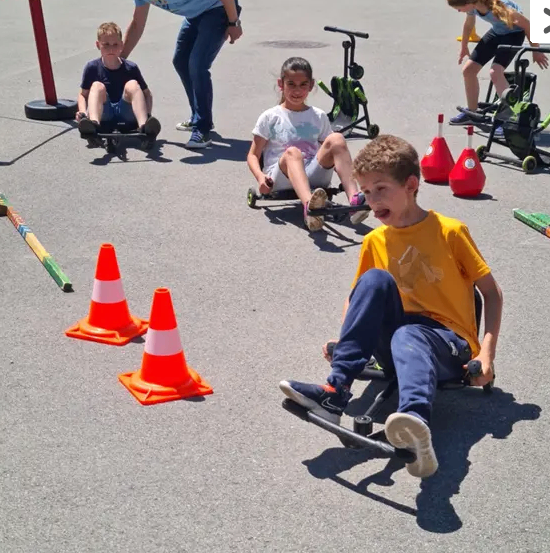 Kinder sitzen auf Go-Karts und auf dem Boden. Einer wird von einem Mann geholfen, andere rasen um Kegel. Im Hintergrund laufen Menschen und es gibt Gebäude.