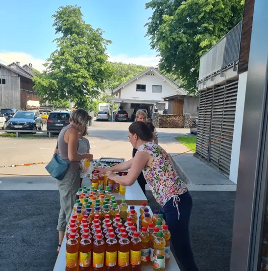 Zwei Frauen bereiten einen Tisch mit Flaschen zum Verkauf vor. Bäume und Häuser sind im Hintergrund.