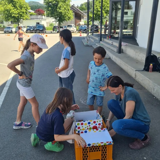 Fünf Kinder und ein Erwachsener versammeln sich um einen orangefarbenen Kasten in einem Außenbereich. Die Kinder stehen, knien und schauen auf den Kasten, während der Erwachsene kniet und lächelt. Im Hintergrund befinden sich Bäume, geparkte Autos und ein Gebäude mit einer Glastür.