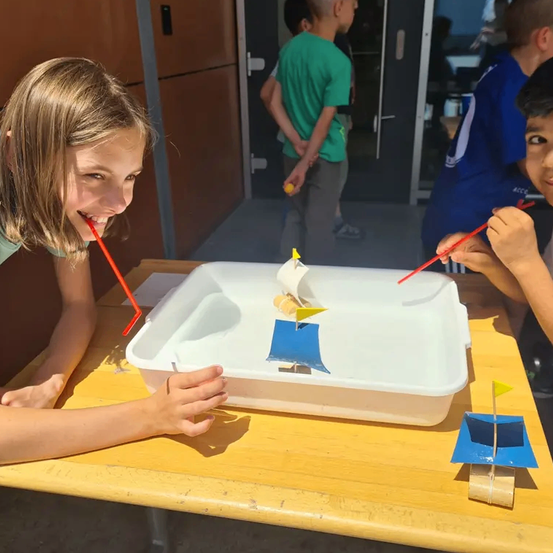 Zwei Kinder spielen mit Spielzeugbooten in einer flachen Wasserwanne. Ein Mädchen bläst auf einen Strohhalm, um ihr Boot zu bewegen. Ein Junge benutzt einen Strohhalm, um sein Boot zu steuern. Sie lächeln, während sie mit den Booten interagieren.