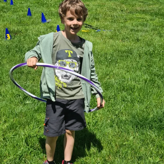 Ein junger Junge hält einen Hula-Hoop auf einem Rasenfeld mit blauen Kegeln im Hintergrund.
