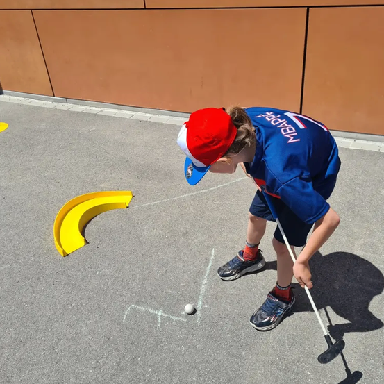 Ein Junge in einem blauen Shirt und rotem Hut übt Golf auf einem gepflasterten Bereich mit einem gelben Hindernis.