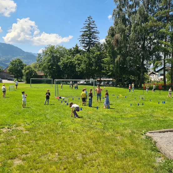 Kinder spielen auf einem Feld mit kleinen Toren und Hütchen. Bäume und Berge sind im Hintergrund.