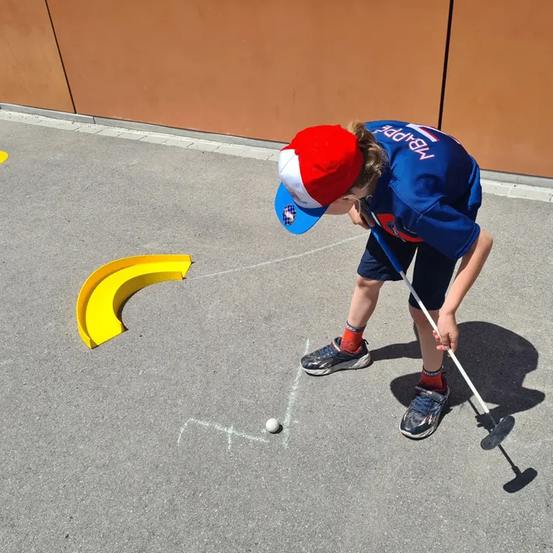 Ein junger Junge mit einer rot-weißen Kappe schlägt einen Golfball mit einem Putter. Der Ball liegt auf dem Boden und der Junge steht in einem schattigen Bereich.
