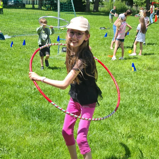 Ein Mädchen in pinkfarbenen Hosen und einer Kappe hält einen Hula-Hoop auf einem Rasenfeld, in der Nähe spielen andere Kinder.