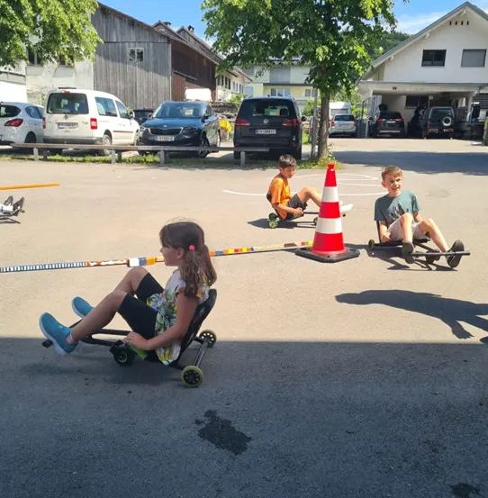 Drei Kinder in Go-Karts rennen auf einer Straße mit einem Verkehrskegel und Gebäuden im Hintergrund.