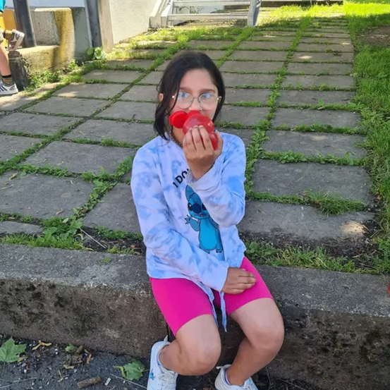 Ein Mädchen mit Brille sitzt auf einem Betonrand, hält einen roten Becher und trägt ein blaues Tie-Dye-Shirt mit einem blauen Cartoon-Charakter und pinke Shorts.