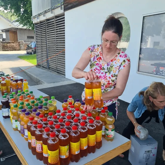 Eine Frau mit Brille hält eine Flasche Saft vor einem Tisch mit vielen Saftflaschen. Eine andere Frau steht neben ihr und schaut sich die Flaschen an.