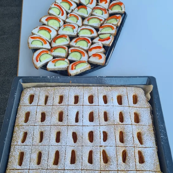 Zwei Tabletts mit Essen stehen auf einem Tisch. Ein Tablett hat ein Gitter aus gebackenen Waren, bestreut mit Puderzucker. Das andere Tablett hat eine Sammlung von Gurkensandwiches, diagonal geschnitten.