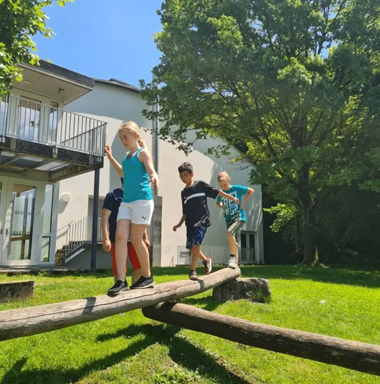 Drei Kinder spielen auf einem Baumstamm-Balancierbalken in einem Grasbereich, mit einem Gebäude und Bäumen im Hintergrund.