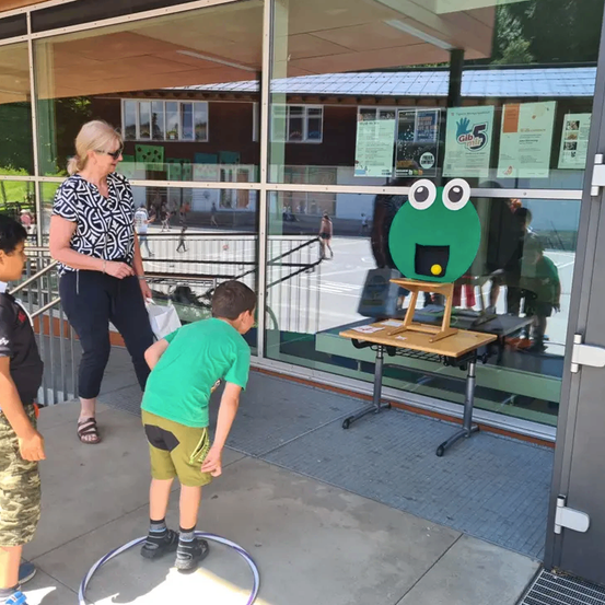 Eine Frau und zwei Jungen stehen vor einer Glastür. Ein Junge beugt sich nach unten, um ein kleines, froschförmiges Objekt auf einem Tisch zu betrachten.