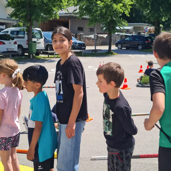 Mehrere Kinder stehen auf einem Parkplatz und beobachten ein Rennen. Sie befinden sich vor einem Gebäude mit geparkten Autos. Verkehrskegel liegen auf dem Boden.