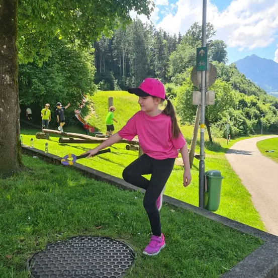 Ein kleines Mädchen in einem rosa T-Shirt und Hut spielt ein Spiel in einem Park mit Grasfläche und Bäumen.
